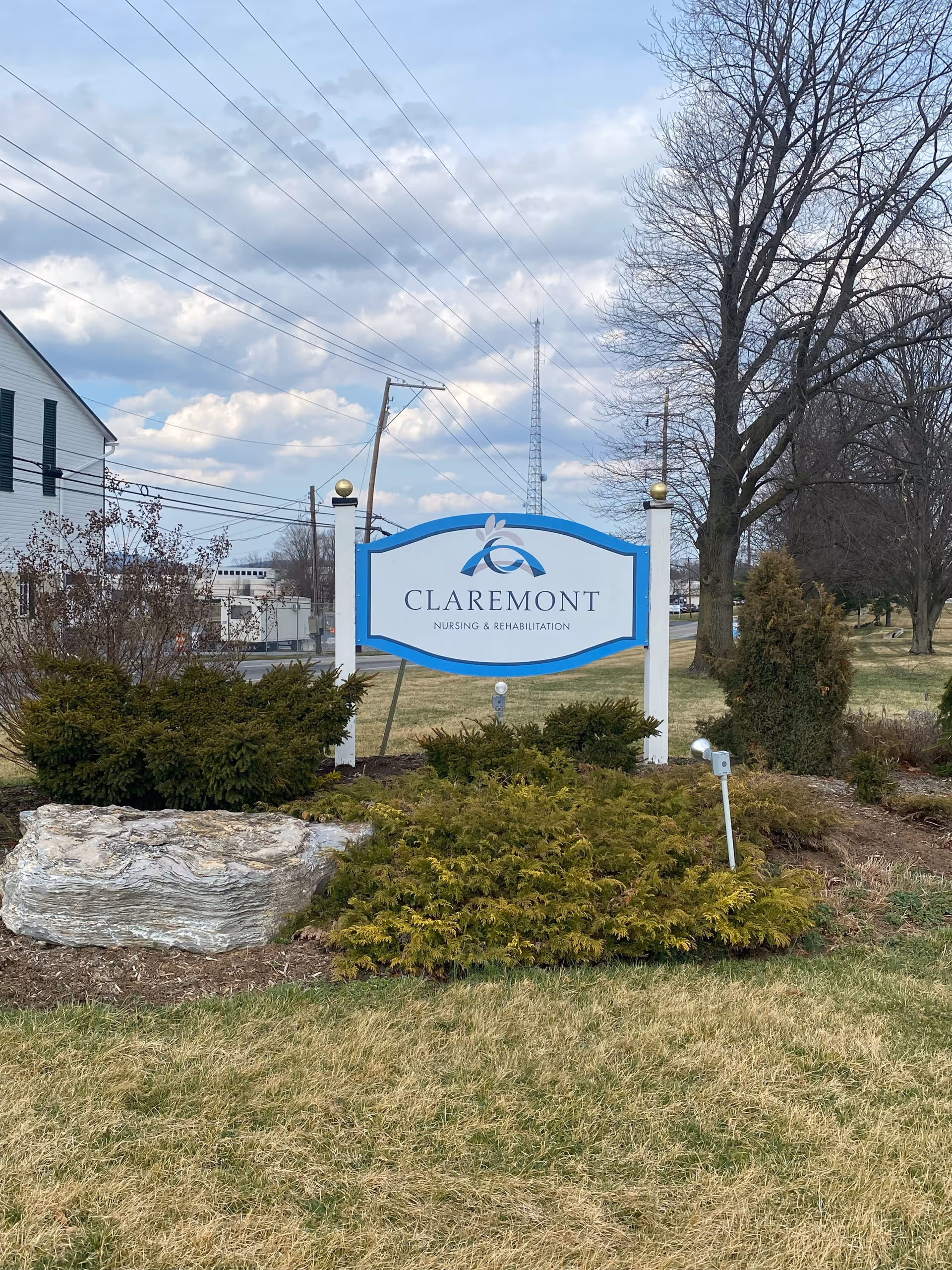 Outdoor view of a sign for Claremont Nursing & Rehabilitation Center surrounded by bushes and grass, with trees and a partly cloudy sky in the background.