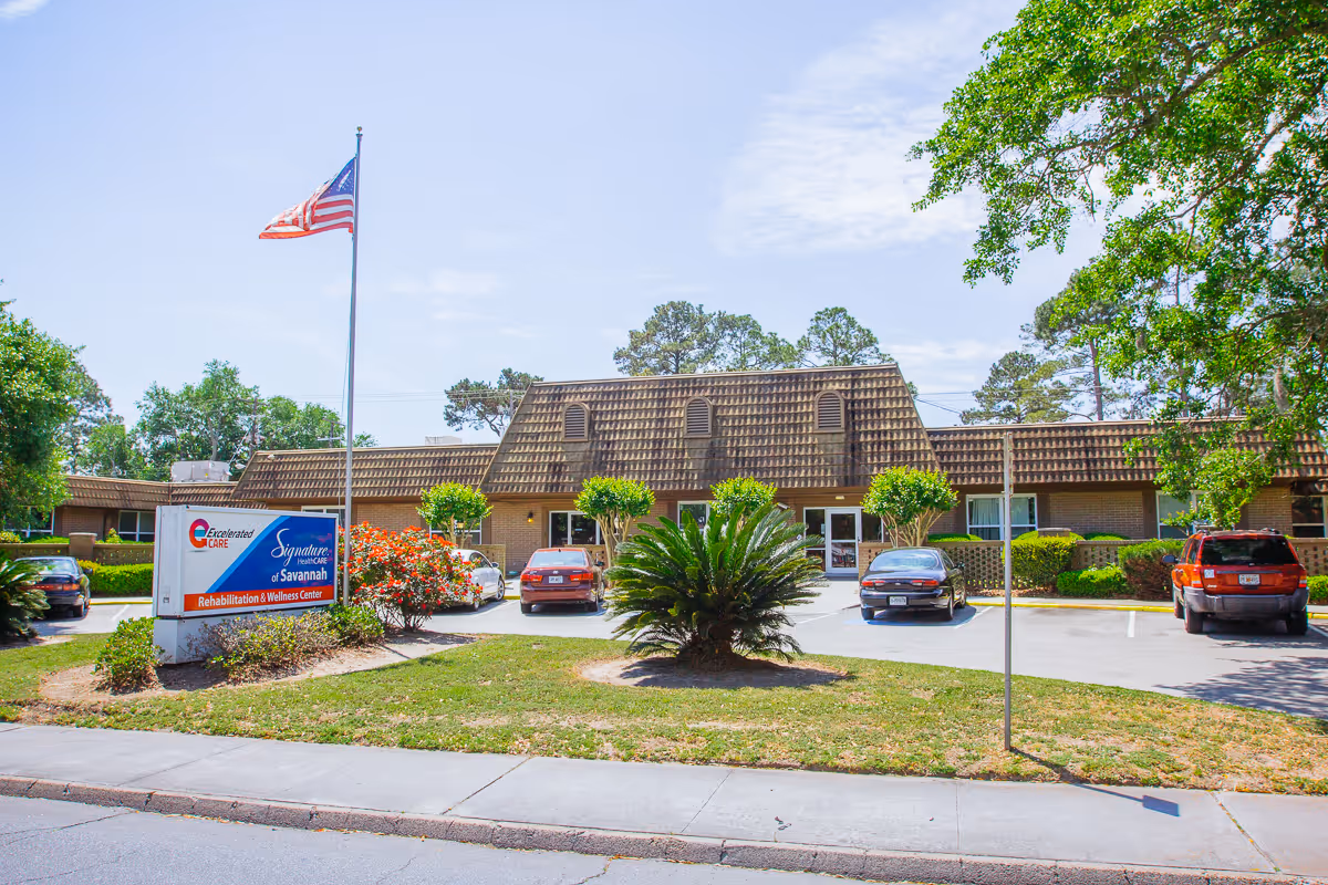Front exterior of a single-story brick senior care facility with a tiled mansard roof, an American flag, landscaped plants, a sign, and several parked cars.