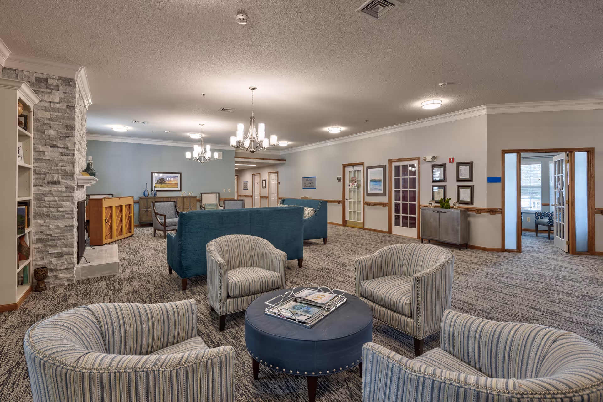 A spacious senior living facility common area with multiple seating arrangements including striped armchairs around a round blue ottoman, teal sofas, and additional chairs near a stone fireplace and piano. The room features carpeted flooring, soft lighting from chandeliers and ceiling fixtures, framed artwork on the walls, and a doorway leading to another room with large windows.