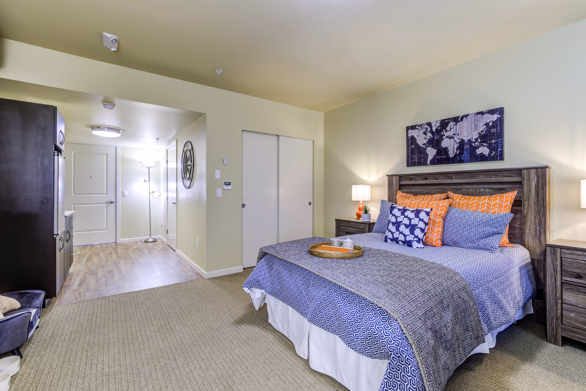 A well-lit bedroom in a senior living facility featuring a large bed with patterned blue and orange pillows and bedding. The room has two wooden nightstands with lamps on either side of the bed. Above the bed hangs a world map artwork. The floor is carpeted, and there is a hallway leading to a door and a closet with sliding doors. A tall floor lamp is visible in the hallway.