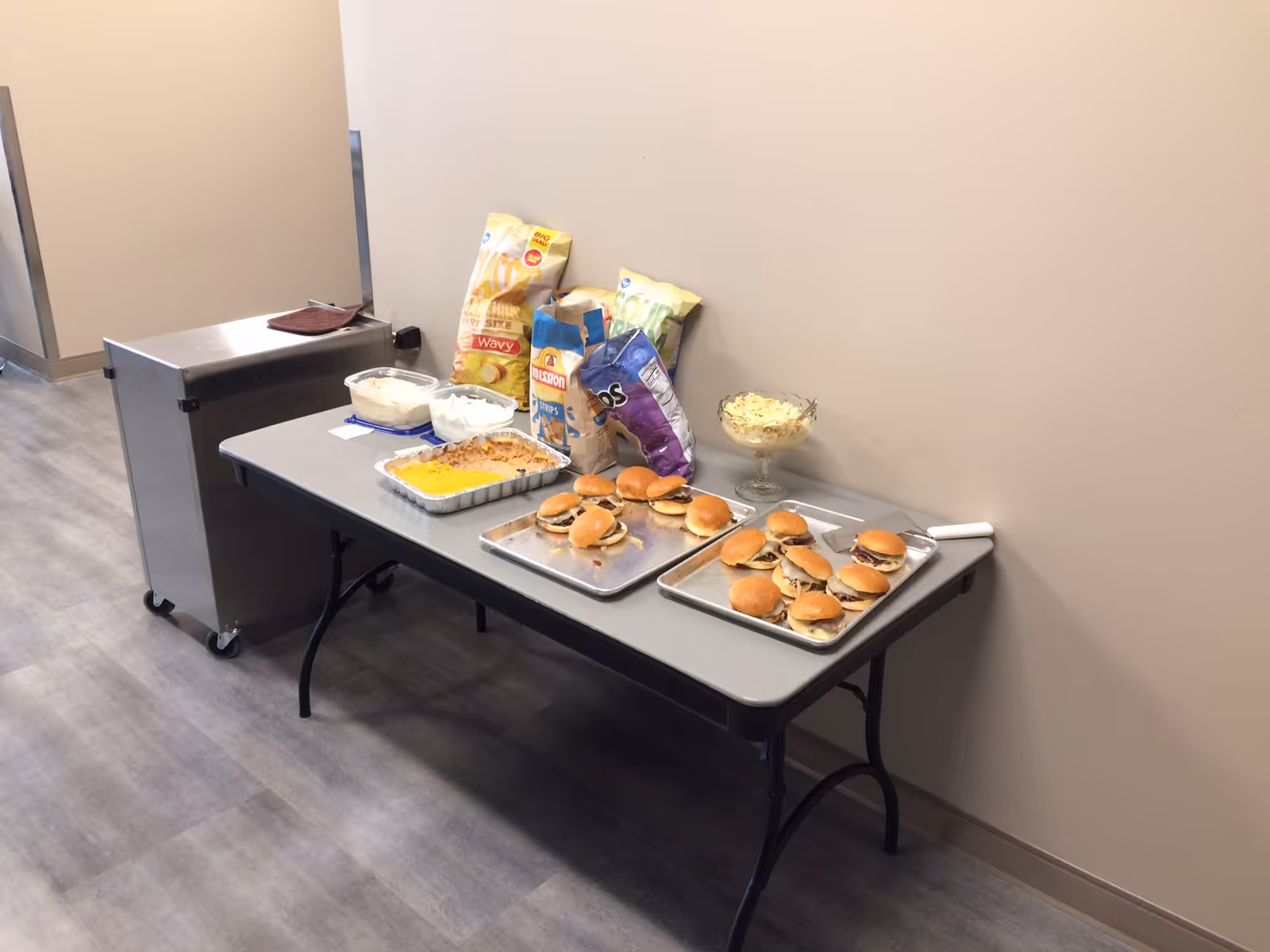 A folding table set against a beige wall with trays of small sandwiches, bags of chips, a casserole dish, and bowls of salad and dip arranged on top. A metal cart with wheels is positioned next to the table on a gray floor.