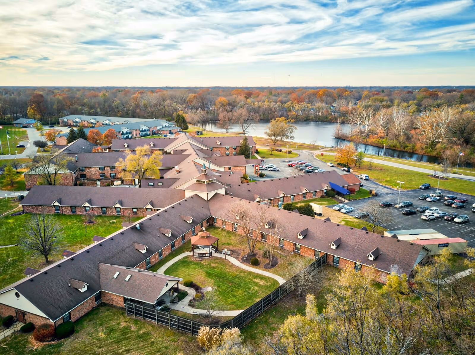 Aerial view of American Village senior living facility showing multiple connected brick buildings with brown roofs, surrounded by green lawns, trees with autumn foliage, parking lots with cars, and a body of water in the background under a partly cloudy sky.