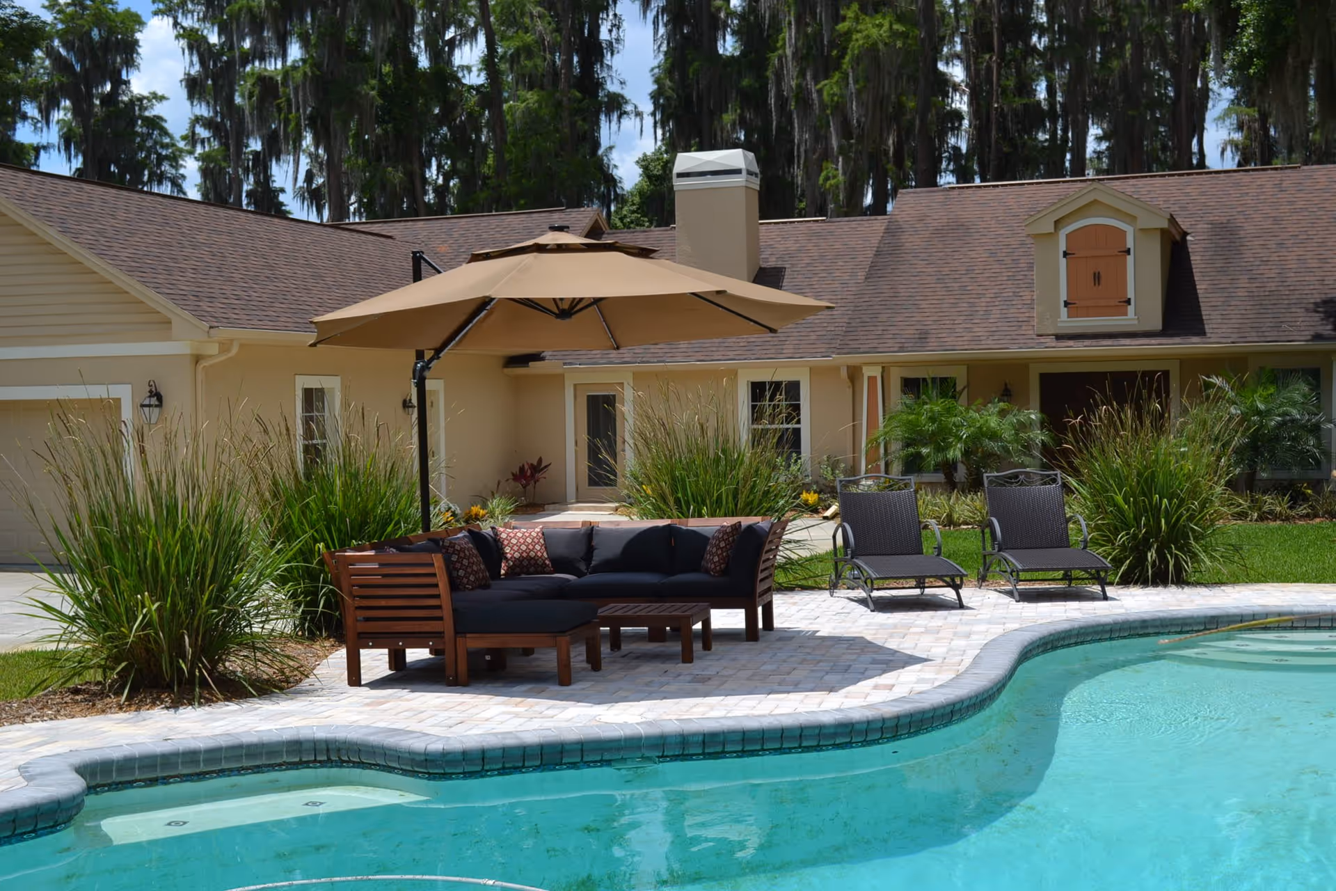 Outdoor patio area at Cloverdale Assisted Living featuring a swimming pool with clear blue water, a wooden sectional sofa with black cushions and patterned pillows under a large beige umbrella, two black lounge chairs, and surrounding greenery including tall grasses and palm plants. The background shows a beige building with a brown shingled roof and trees with hanging moss.
