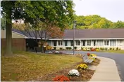 Exterior view of a single-story senior living facility building with a brown roof, surrounded by green trees and a landscaped area with colorful flowers along a curved sidewalk.