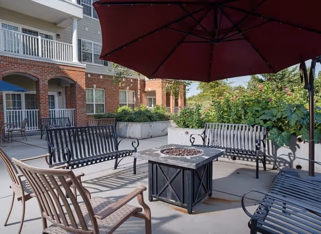 Outdoor patio area at Harmony at Chantilly with metal benches and chairs arranged around a square fire pit table, shaded by a large red umbrella. The background shows a brick building with balconies and greenery.