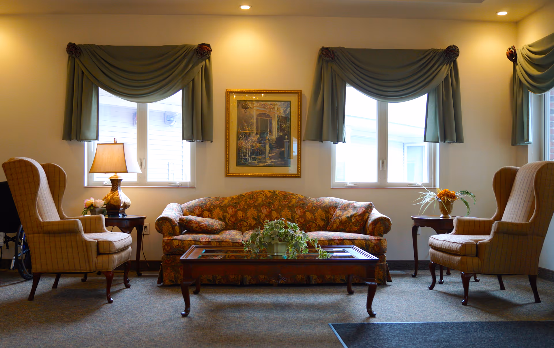 A cozy living room area with a floral patterned sofa centered between two large windows with green drapes. On either side of the sofa are beige wingback chairs with a small wooden side table next to each, one holding a lamp and the other a flower arrangement. A wooden coffee table with a glass top and a green potted plant sits in front of the sofa. The walls are light-colored and there is a framed painting hanging between the windows.
