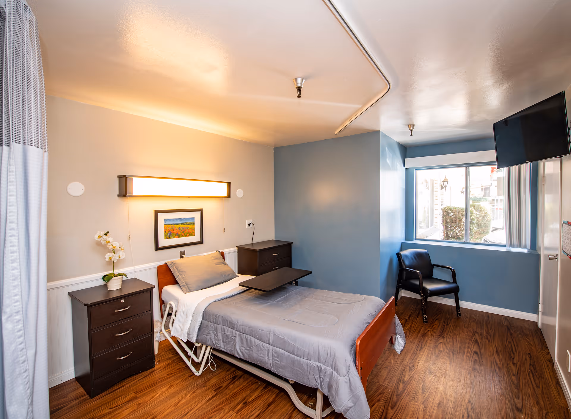A small, clean hospital room with a single bed covered in gray bedding. The bed has an adjustable overbed table. There are two dark wooden nightstands, one with a potted orchid. A framed picture hangs on the wall above the bed, which is illuminated by a long wall light. A black chair is positioned near a window with vertical blinds, and a flat-screen TV is mounted on the wall. The floor is wood laminate, and the walls are painted light gray and blue.