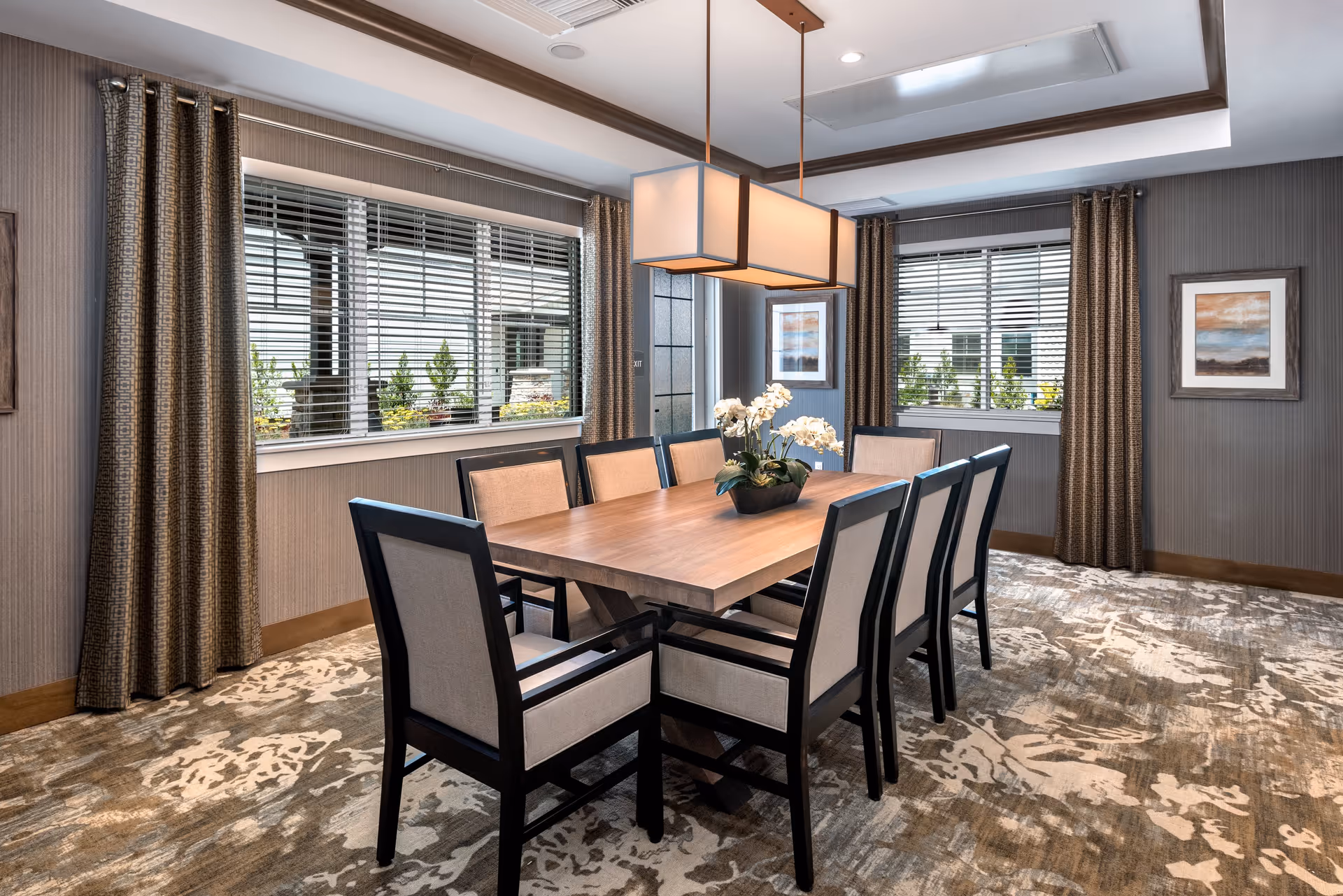 A dining room with a rectangular wooden table surrounded by eight cushioned chairs with dark frames. The room has two large windows with blinds and patterned curtains. A modern rectangular pendant light hangs above the table, and there is a floral centerpiece on the table. The walls are decorated with framed artwork, and the floor is covered with a patterned carpet.