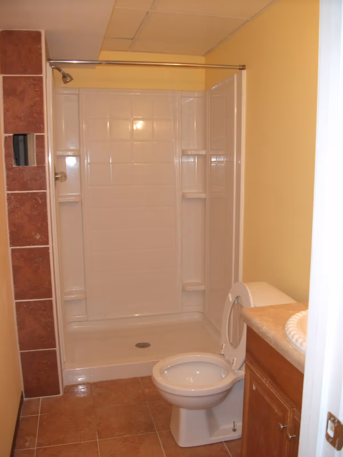 A small bathroom with a white toilet with the lid open, a wooden vanity with a beige countertop and a white sink, and a white tiled shower with a metal showerhead and a metal shower curtain rod. The walls are painted yellow and the floor is covered with brown tiles. There is a section of wall with brown tiles next to the shower.