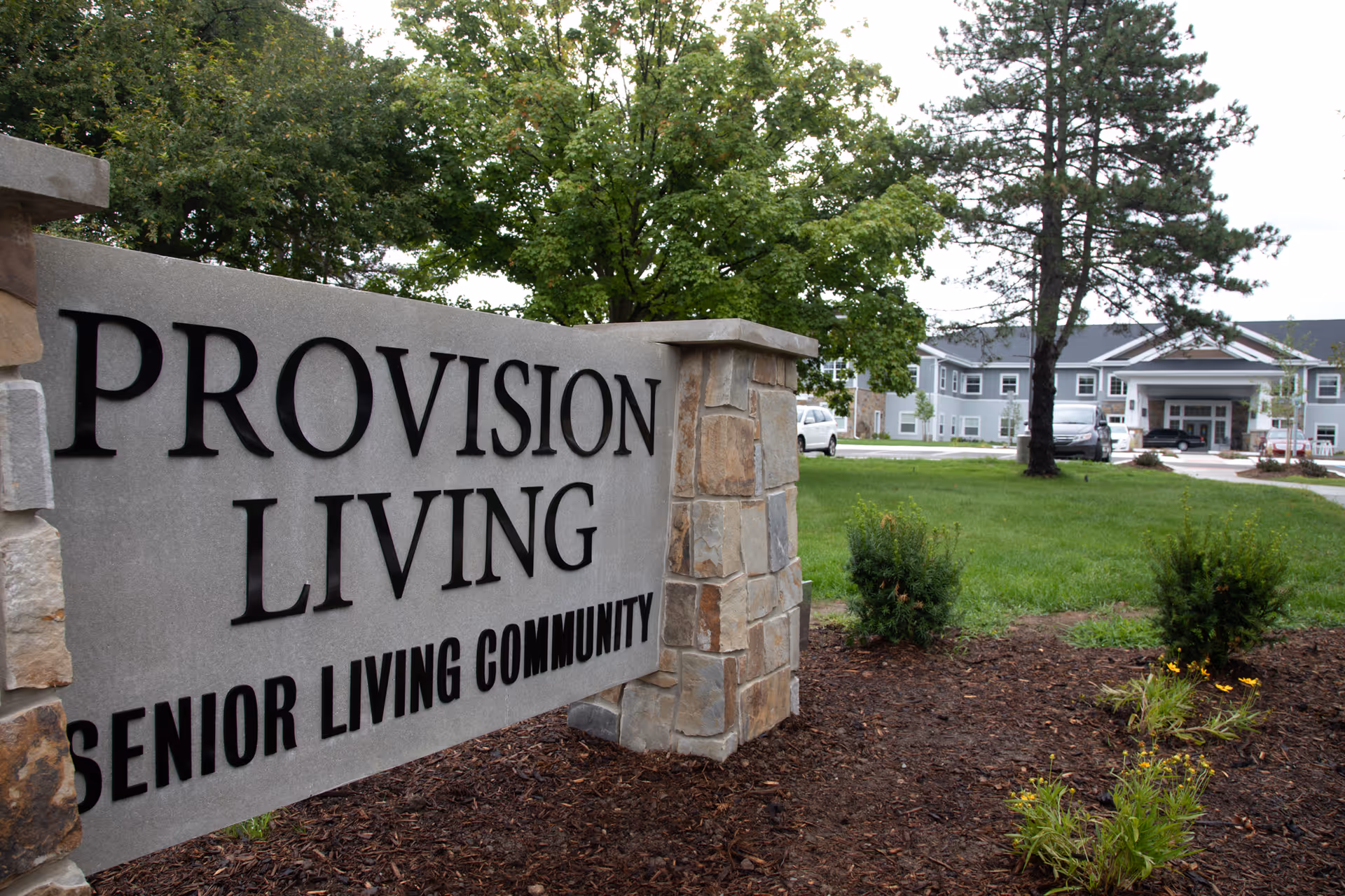 A stone sign reading 'PROVISION LIVING SENIOR LIVING COMMUNITY' in front of a lawn with the facility building and entrance visible in the background.