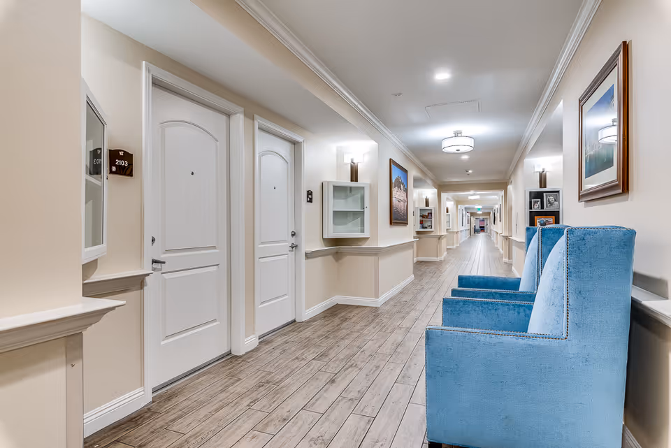 Long, well-lit interior hallway with white apartment doors and blue upholstered chairs along the wall.