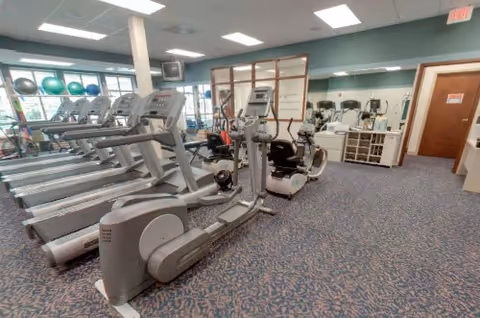 Interior view of a fitness room with several treadmills, elliptical machines, and stationary bikes. The room has blue patterned carpet, light blue walls, large windows letting in natural light, and exercise balls on a rack near the windows.