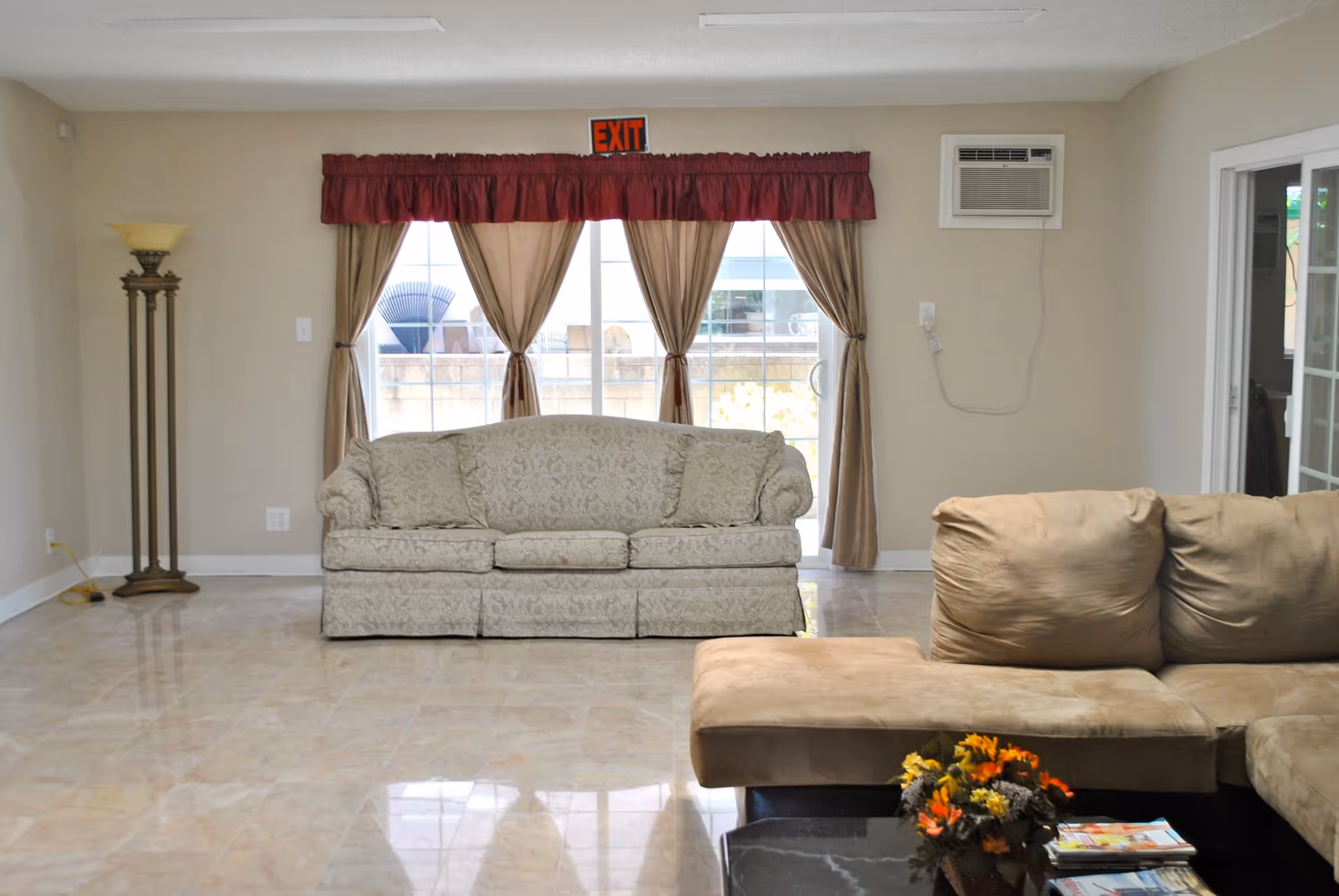 A living room with a beige patterned sofa against a wall with large windows covered by beige curtains and a red valance. There is a beige sectional sofa in the foreground with a black coffee table holding a flower arrangement and magazines. A tall floor lamp stands in the corner, and an air conditioning unit is mounted on the wall.