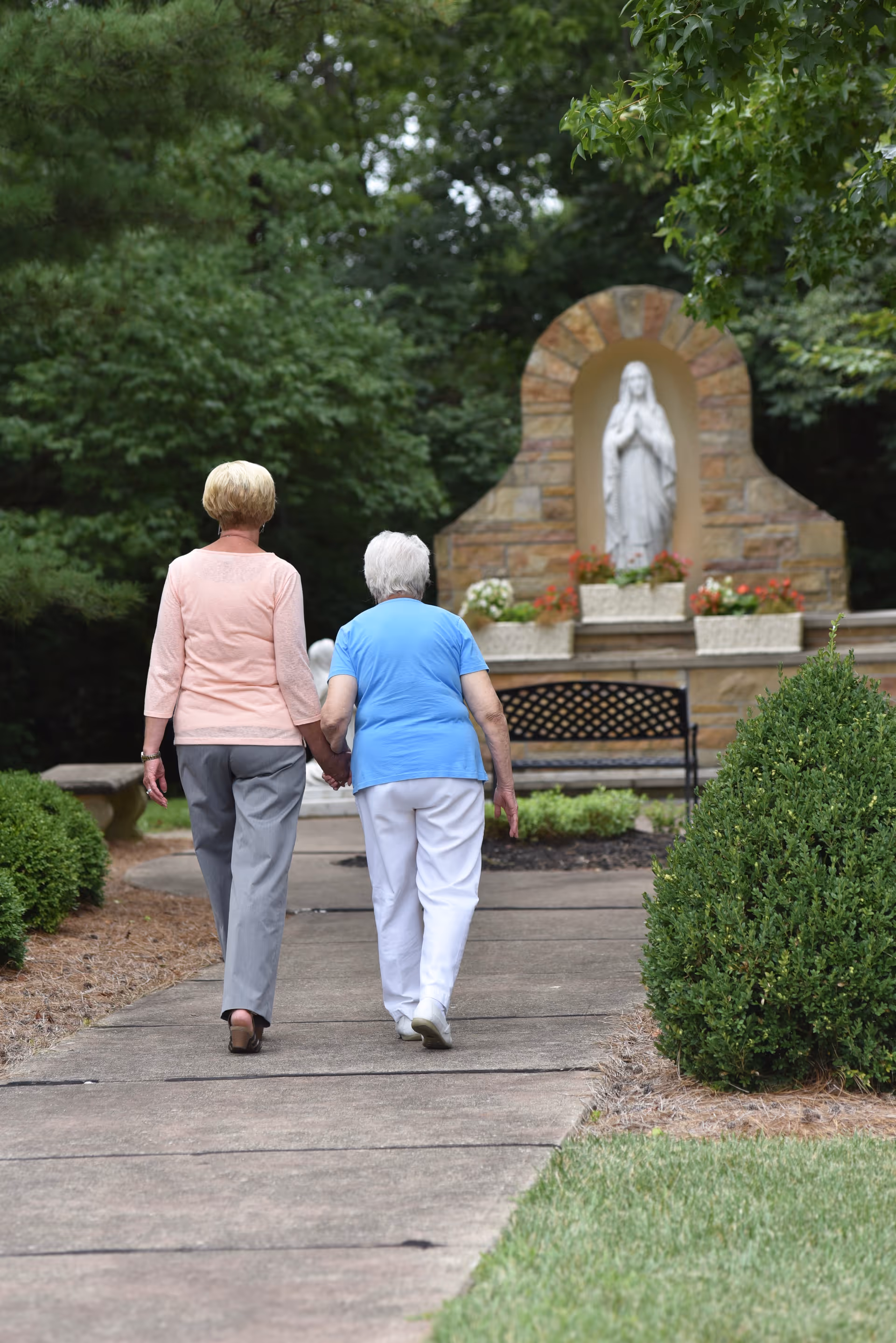 Two elderly women walking hand in hand on a paved path in a garden area with green bushes and trees. In the background, there is a stone shrine with a statue of a praying figure and flower pots.