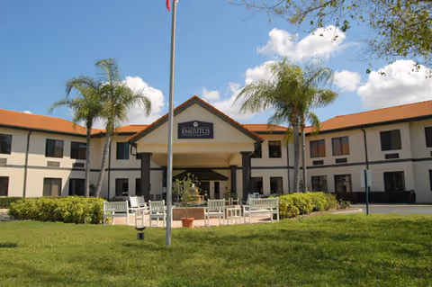 Front exterior of a two-story senior living facility with a covered entrance, palm trees, flagpole, and outdoor seating on a grassy lawn.