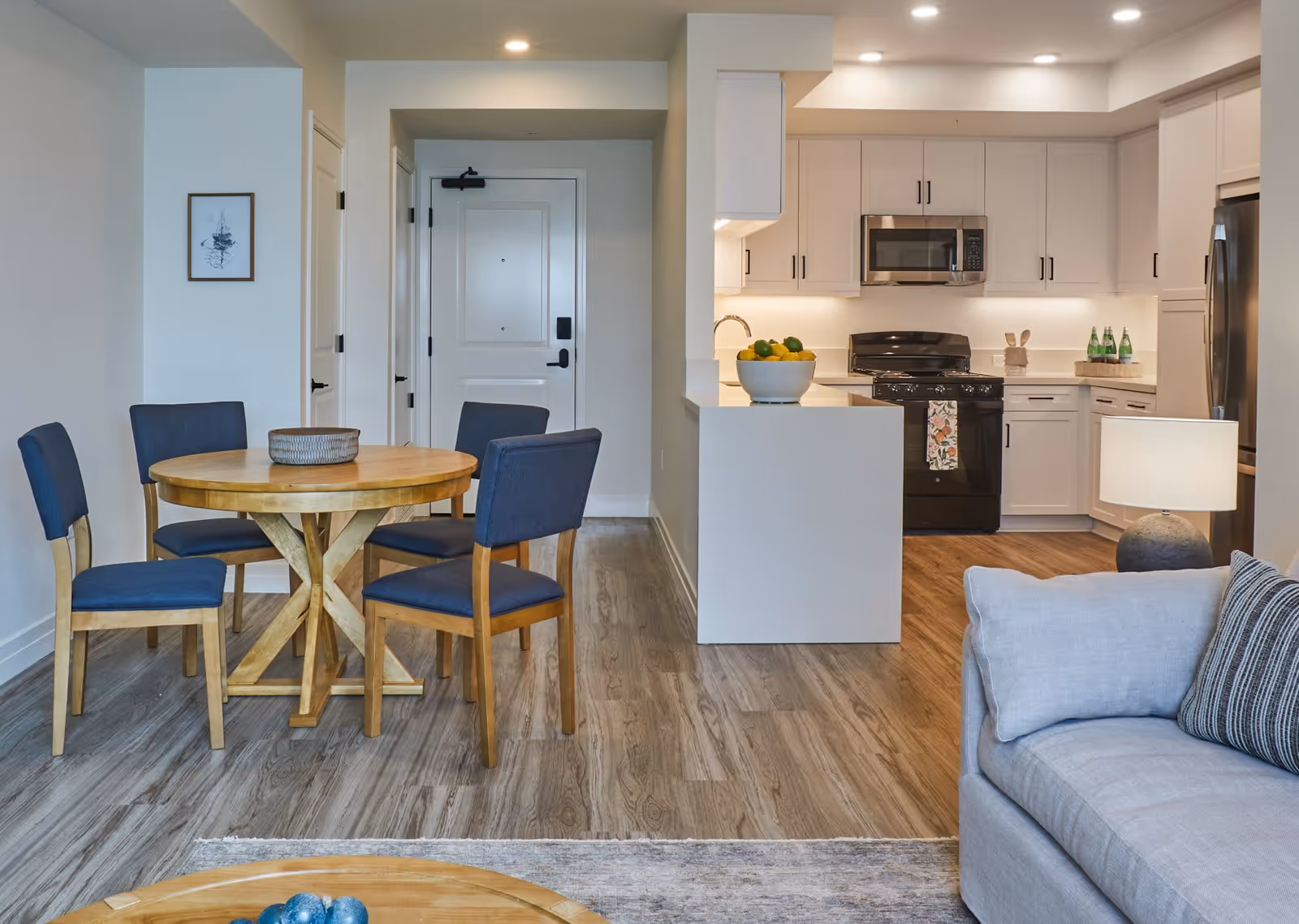 Interior view of a senior living facility showing a dining area with a round wooden table and four blue cushioned chairs, adjacent to a modern kitchen with white cabinets, a black stove, microwave, and refrigerator. A light gray couch with a striped pillow and a lamp on a side table are partially visible in the foreground.