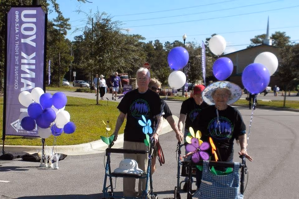 A group of elderly people walking outdoors on a sunny day, some using walkers decorated with colorful pinwheels and balloons. They are wearing matching black t-shirts with a logo, and one woman is wearing a wide-brimmed hat. Purple and white balloons and a purple banner with the words 'THANK YOU' are visible in the background.