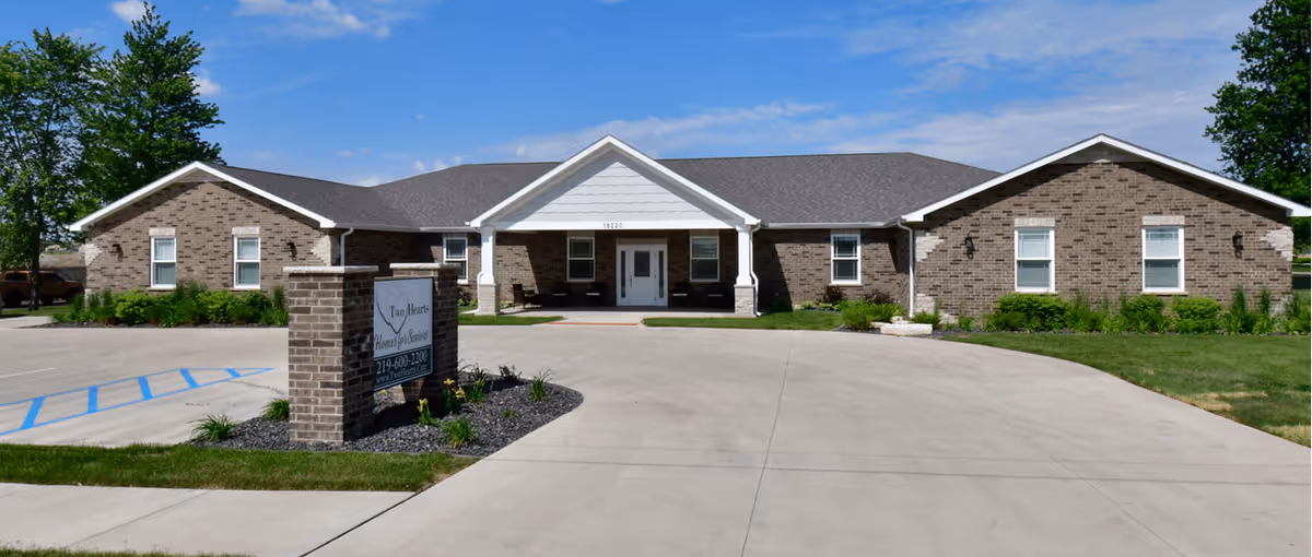 Front exterior view of a single-story brick building with a gray roof and white trim, featuring a covered entrance with seating. There is a paved driveway and parking area with a sign in front that reads 'Two Hearts Homes For Seniors'. The sky is clear and blue with some trees in the background.