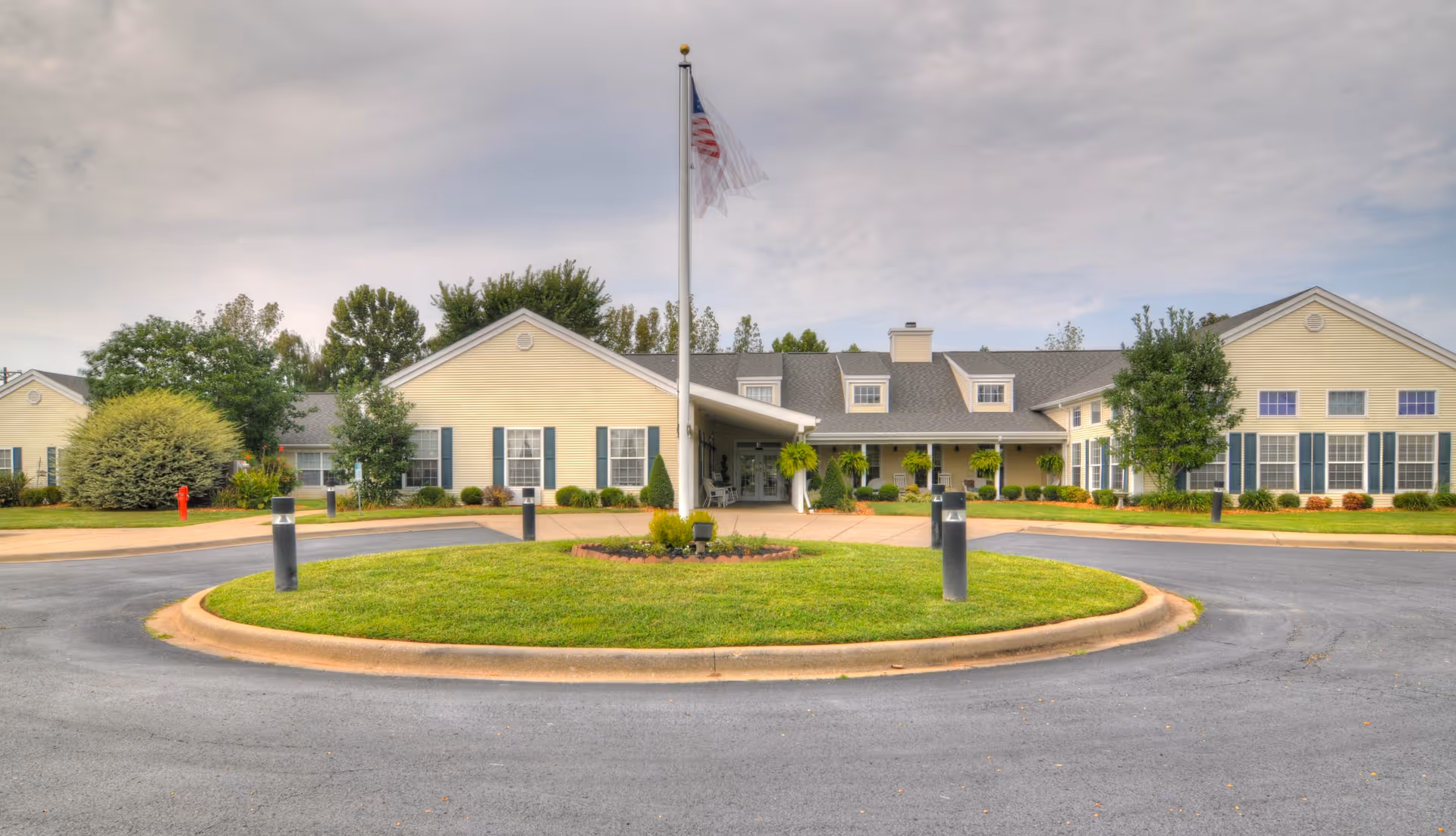 Front exterior view of a single-story senior living facility building with a circular driveway and a flagpole with an American flag in the center. The building is light yellow with white trim and has several windows with green shutters. There are trees and shrubs around the building and a cloudy sky overhead.