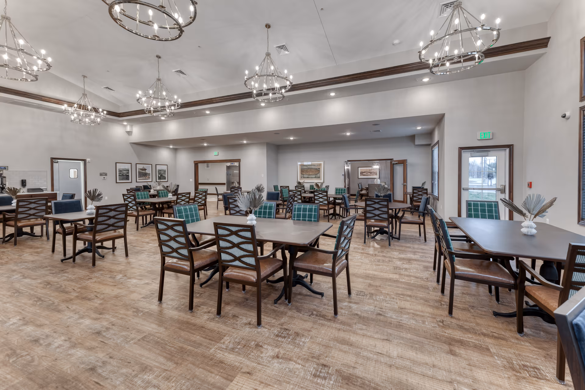 A spacious senior living dining room with multiple tables and chairs arranged neatly. The room features wooden flooring, modern chandeliers hanging from a high ceiling, and framed artwork on the walls. There are exit doors and windows allowing natural light to enter.