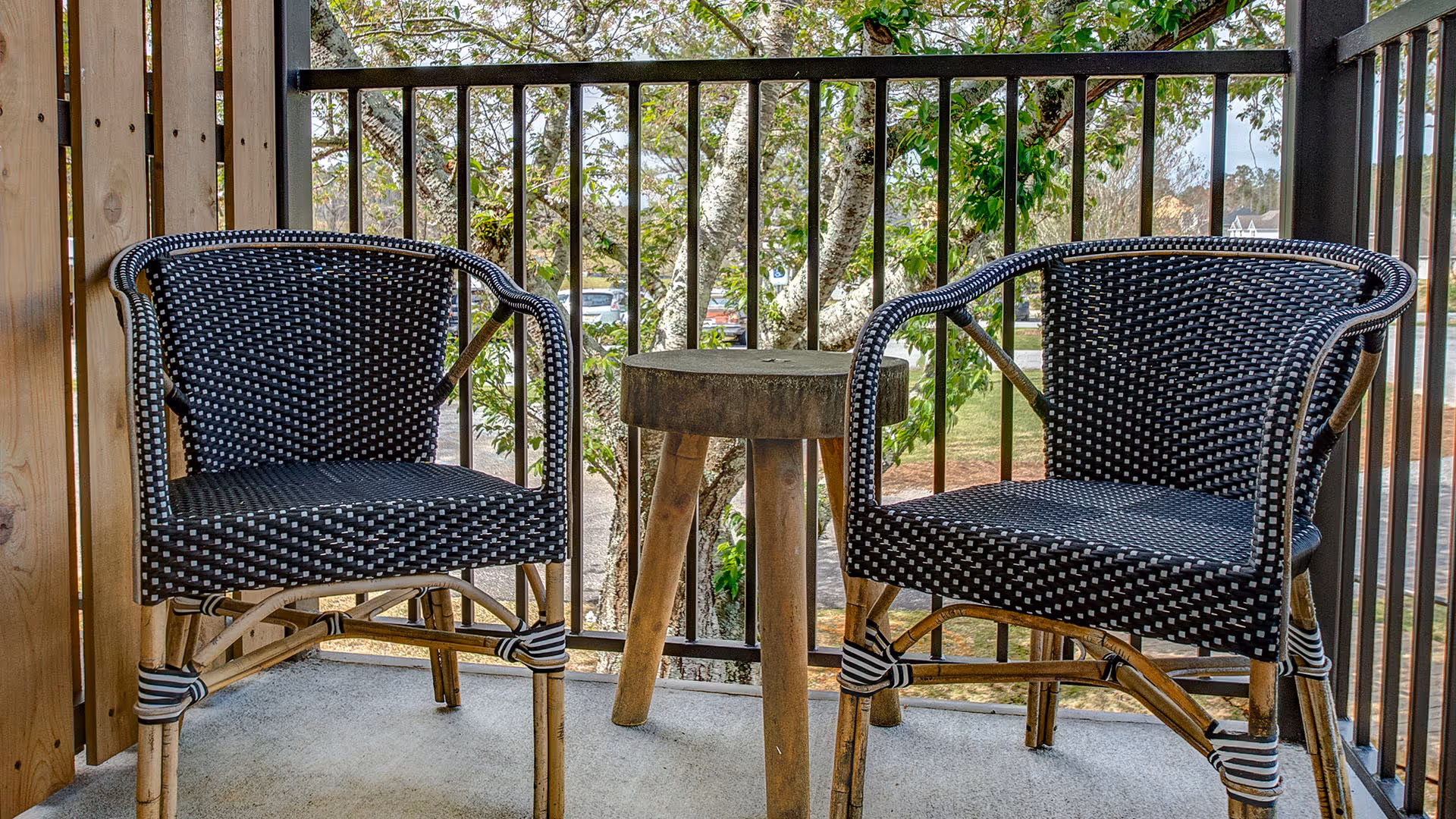 Two black and white woven chairs with wooden frames and a small wooden round table between them on a balcony with black metal railings and a tree with green leaves in the background.