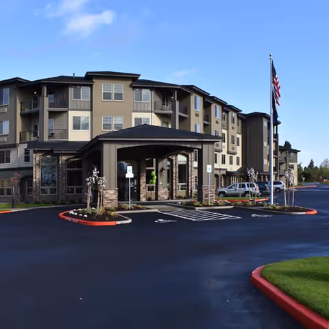 Exterior view of a multi-story senior living facility building with a covered entrance, several windows, a flagpole with an American flag, and a parking area with marked handicap spaces.