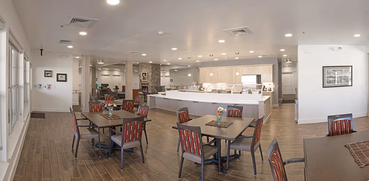 A spacious dining area in a senior living facility with several tables and chairs arranged neatly. The room features wood-look flooring, large windows on the left side, and a kitchen area with a long counter and modern appliances in the background. The walls are decorated with framed pictures, and the ceiling has recessed lighting.