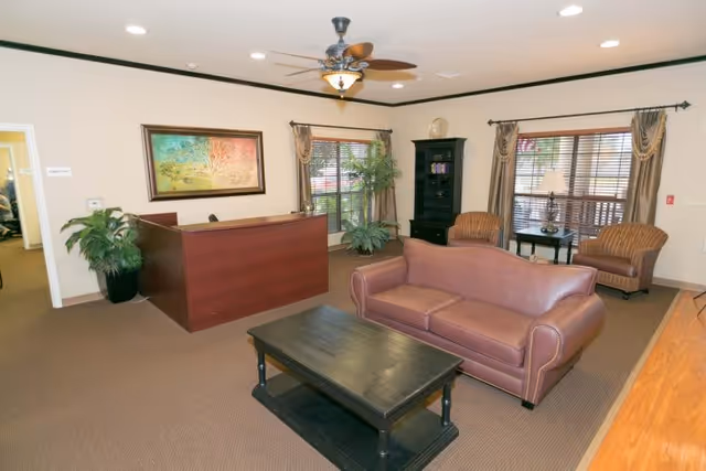 Reception-style lounge with a leather sofa, coffee table, reception desk, armchairs, plants, and windows.