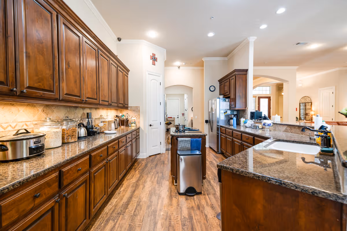 A spacious kitchen with wooden cabinets and granite countertops. The kitchen features a long counter with various appliances and containers, a central island with a trash bin, and a refrigerator. The floor is wooden, and the room is well-lit with recessed lighting. The kitchen opens into a living area with visible doorways and archways.
