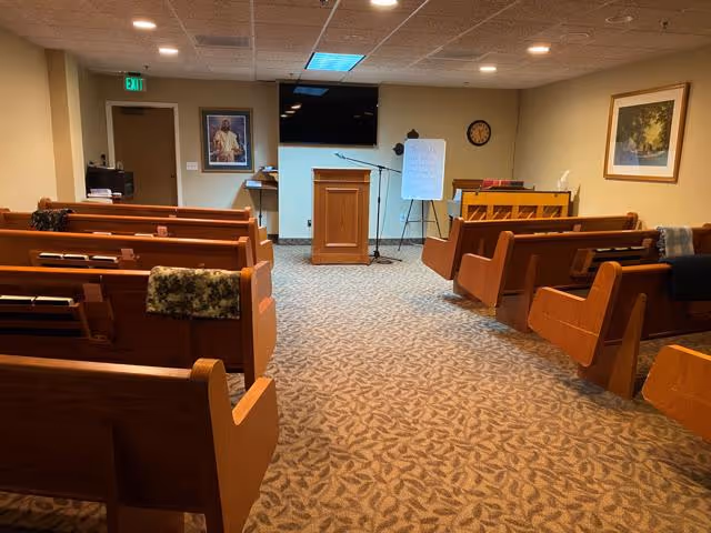 Small chapel-style meeting room with wooden pews facing a podium, flat-screen TV, and a whiteboard.