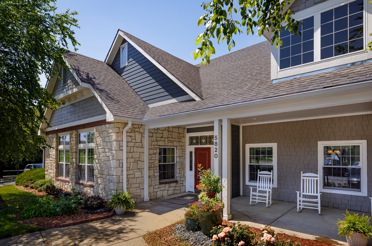 Front entrance of a senior living building with a covered porch, rocking chairs, stone facade, and landscaped walkway.