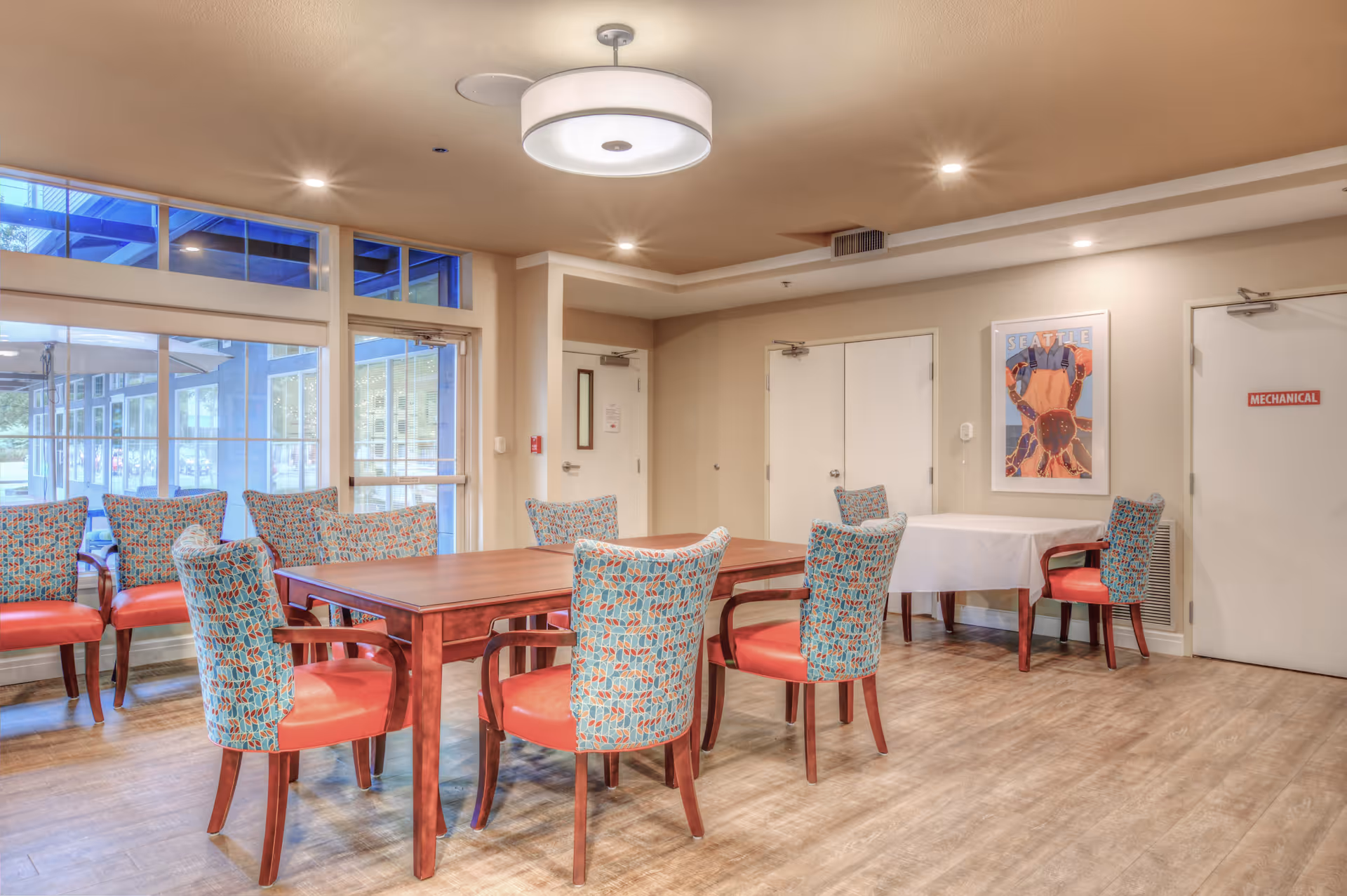 A bright dining area with a wooden table surrounded by eight chairs upholstered in a colorful patterned fabric with red seats. There is a smaller table covered with a white tablecloth against the wall with two additional chairs. The room has large windows and glass doors letting in natural light, light-colored walls, and a ceiling light fixture. A framed artwork is hung on the wall, and there are two doors, one labeled 'MECHANICAL'.