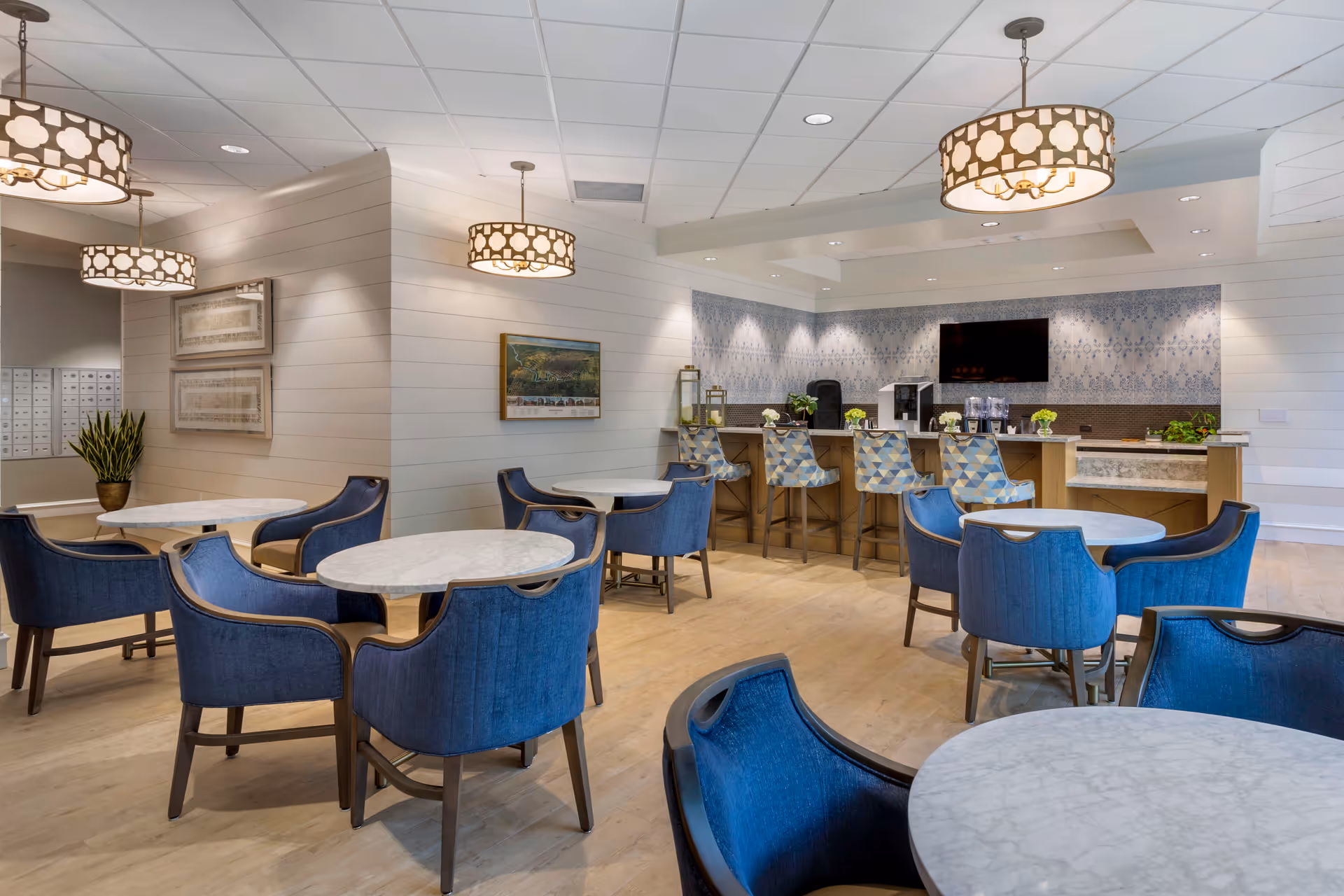 A modern dining area with round marble tables and blue upholstered chairs. There is a counter with patterned bar stools and a coffee station against a wall with decorative blue and white tiles. Three pendant lights with patterned shades hang from the ceiling, and there are framed pictures and a plant on the walls.