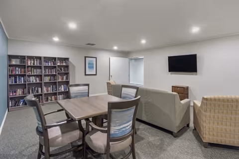 A cozy common area with a wooden table and four chairs in the foreground, two sofas and an armchair facing a wall-mounted flat screen TV, and a bookshelf filled with books against the wall. The room has neutral-colored walls and carpeted flooring with recessed ceiling lights.
