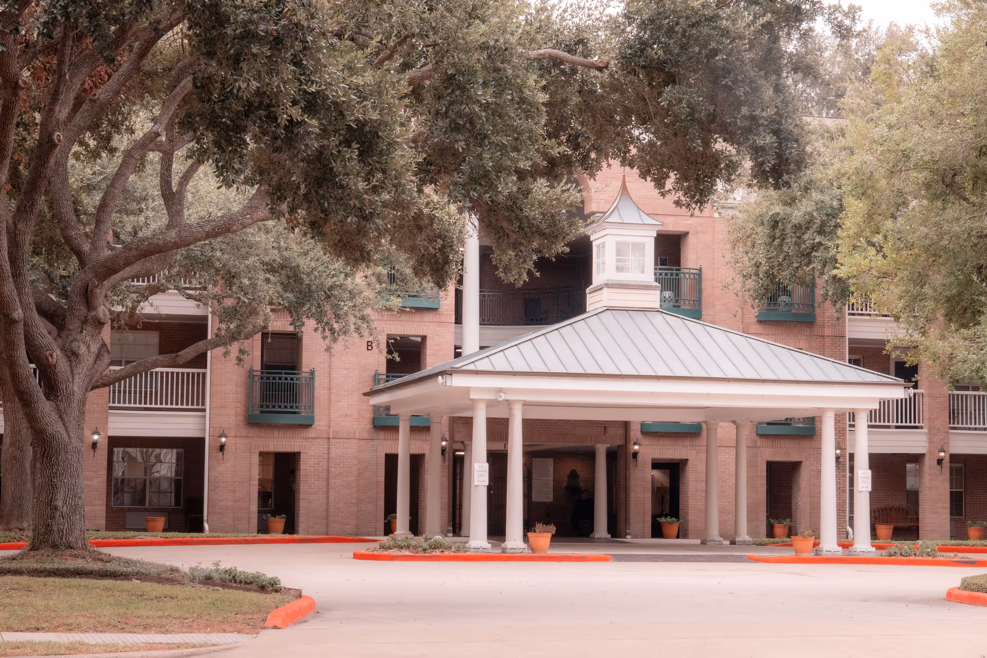 Entrance of a senior living facility with a covered driveway supported by white columns, surrounded by large trees and a brick building with balconies in the background.