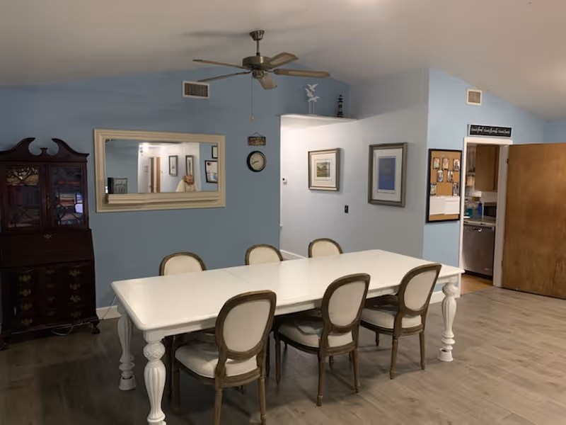 A dining area with a long white table surrounded by six upholstered chairs with wooden frames. The walls are painted light blue and decorated with framed pictures and a clock. A large mirror reflects part of the room, and a wooden cabinet stands against one wall. The floor is light wood, and a doorway leads to a kitchen area with visible appliances.