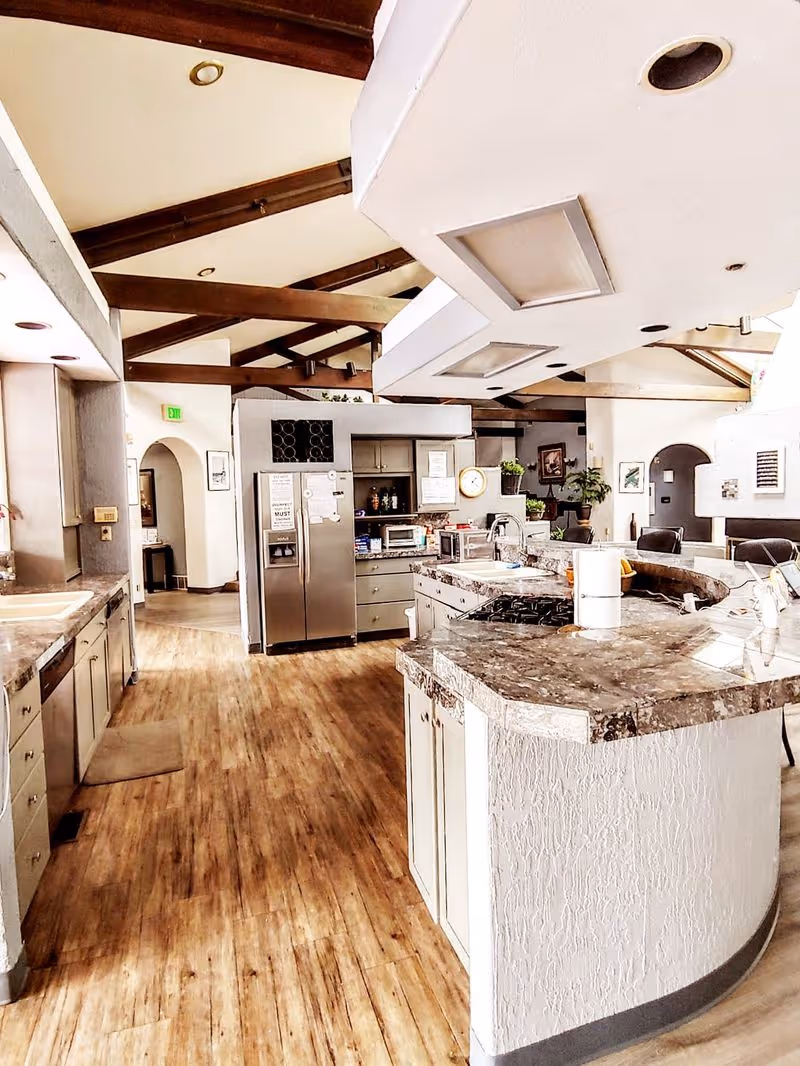Spacious kitchen area with wooden floors, a large curved marble countertop island with a stove, stainless steel refrigerator, and cabinets. The ceiling has exposed wooden beams and recessed lighting. There are plants and framed pictures in the background, and an arched doorway leading to another room.