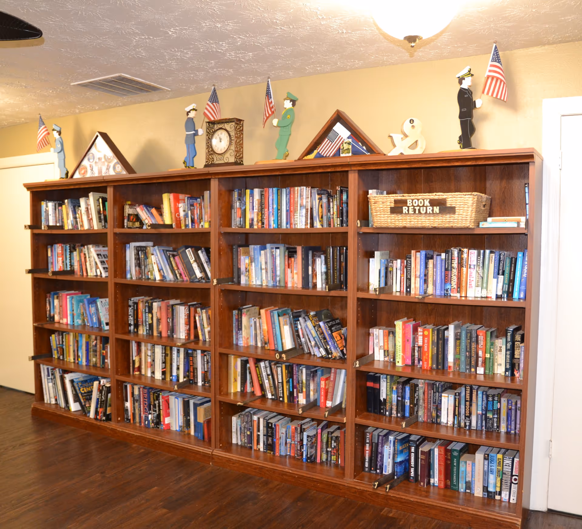 A large wooden bookshelf filled with various books in a senior living community. On top of the bookshelf are decorative figurines of military personnel holding American flags, a clock, and a basket labeled 'BOOK RETURN'. The room has a wooden floor and beige walls.