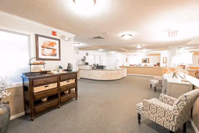 A bright and spacious interior common area with beige walls and carpeted floor. There is a dark wooden sideboard with baskets and a coffee setup on the left, two patterned armchairs on the right, and a curved reception desk in the background. The area is well-lit with ceiling lights and has framed artwork on the walls.