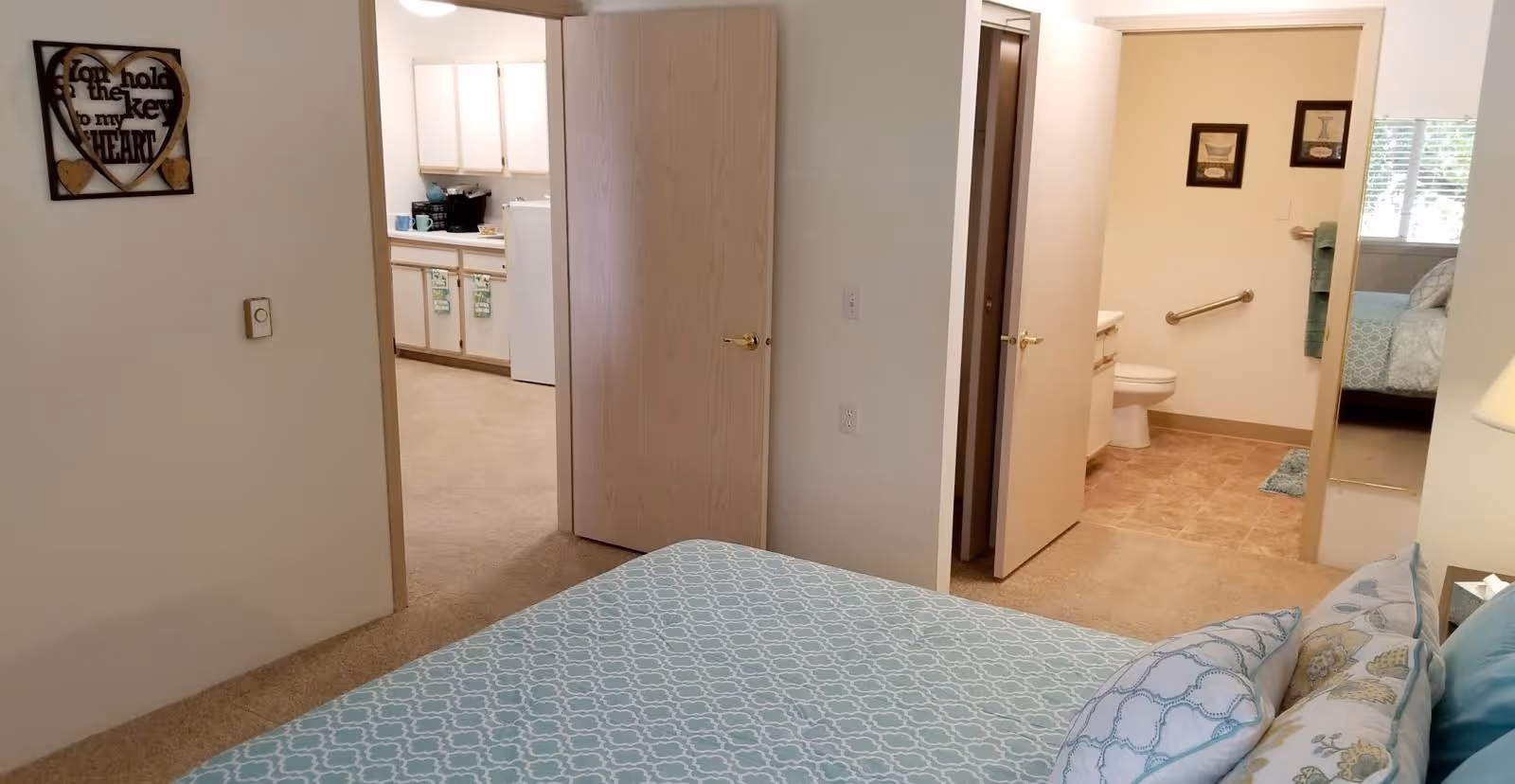 View from a bedroom showing a bed with patterned blue and white bedding, an open door leading to a kitchen with white cabinets and appliances, and another open door leading to a bathroom with a toilet, grab bar, and framed pictures on the wall.