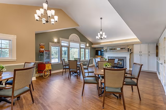 A spacious and well-lit dining area with multiple round tables and chairs arranged neatly. The room features large windows allowing natural light to fill the space, a popcorn machine in the corner, two modern chandeliers hanging from the ceiling, and a fireplace with a TV mounted above it. The walls are painted in soft beige and green tones, and the floor is covered with wood-style flooring.
