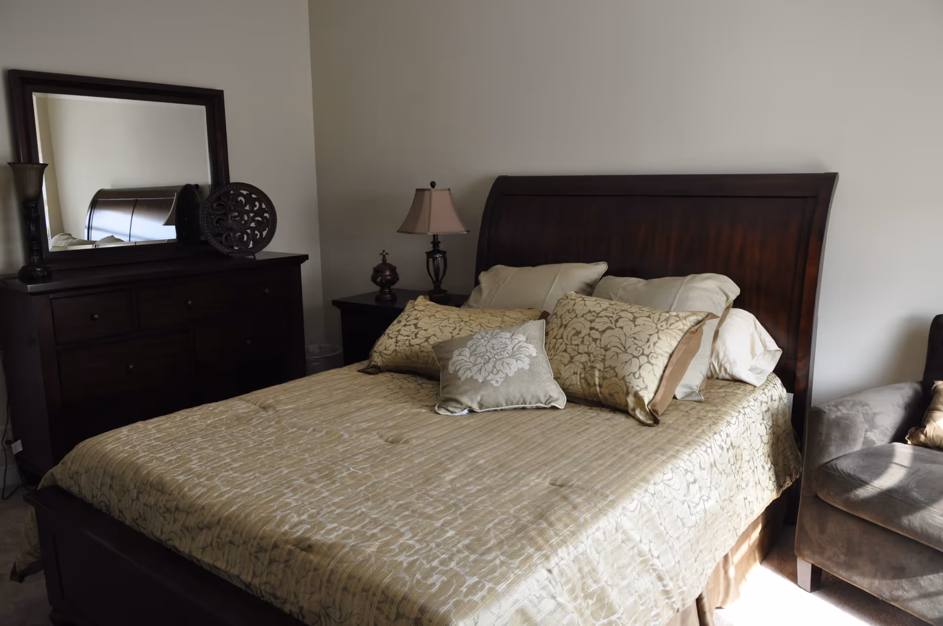 A bedroom with a large wooden bed featuring a beige patterned bedspread and multiple pillows. To the left of the bed is a dark wooden dresser with a large mirror and decorative items on top. A bedside table with a lamp and a small decorative object is next to the bed. On the right side, there is a gray upholstered armchair partially visible. The walls are plain and light-colored.