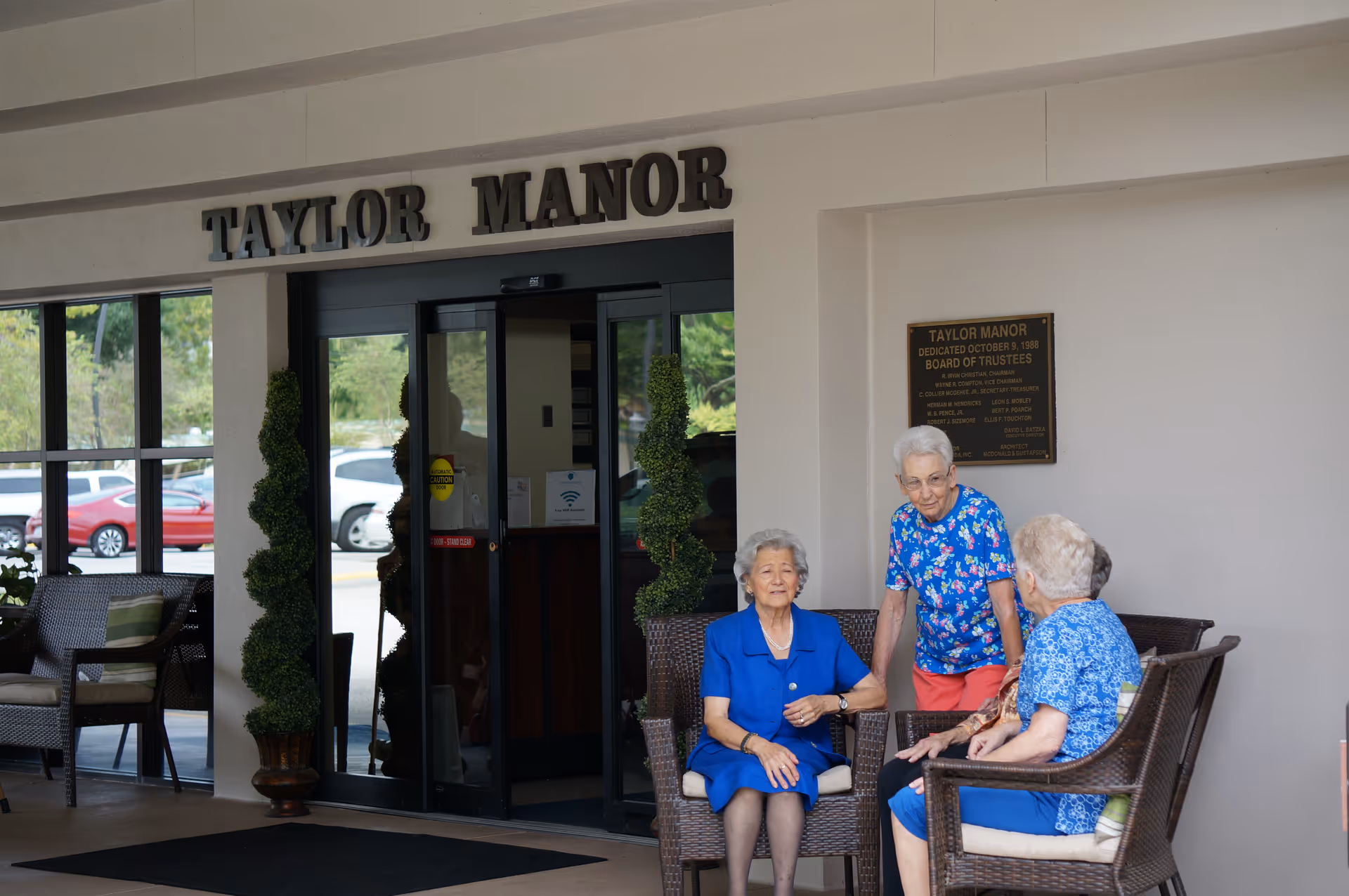 Three elderly women sit and chat in wicker chairs outside the glass entrance of Taylor Manor beneath the building's sign.