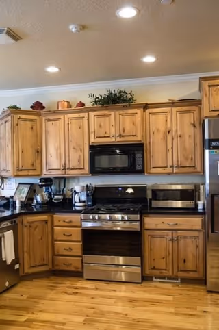 A kitchen with wooden cabinets, a stainless steel oven and stove, a microwave above the stove, a toaster oven on the counter, and various kitchen appliances on the countertop. The floor is wooden and there are recessed lights in the ceiling.