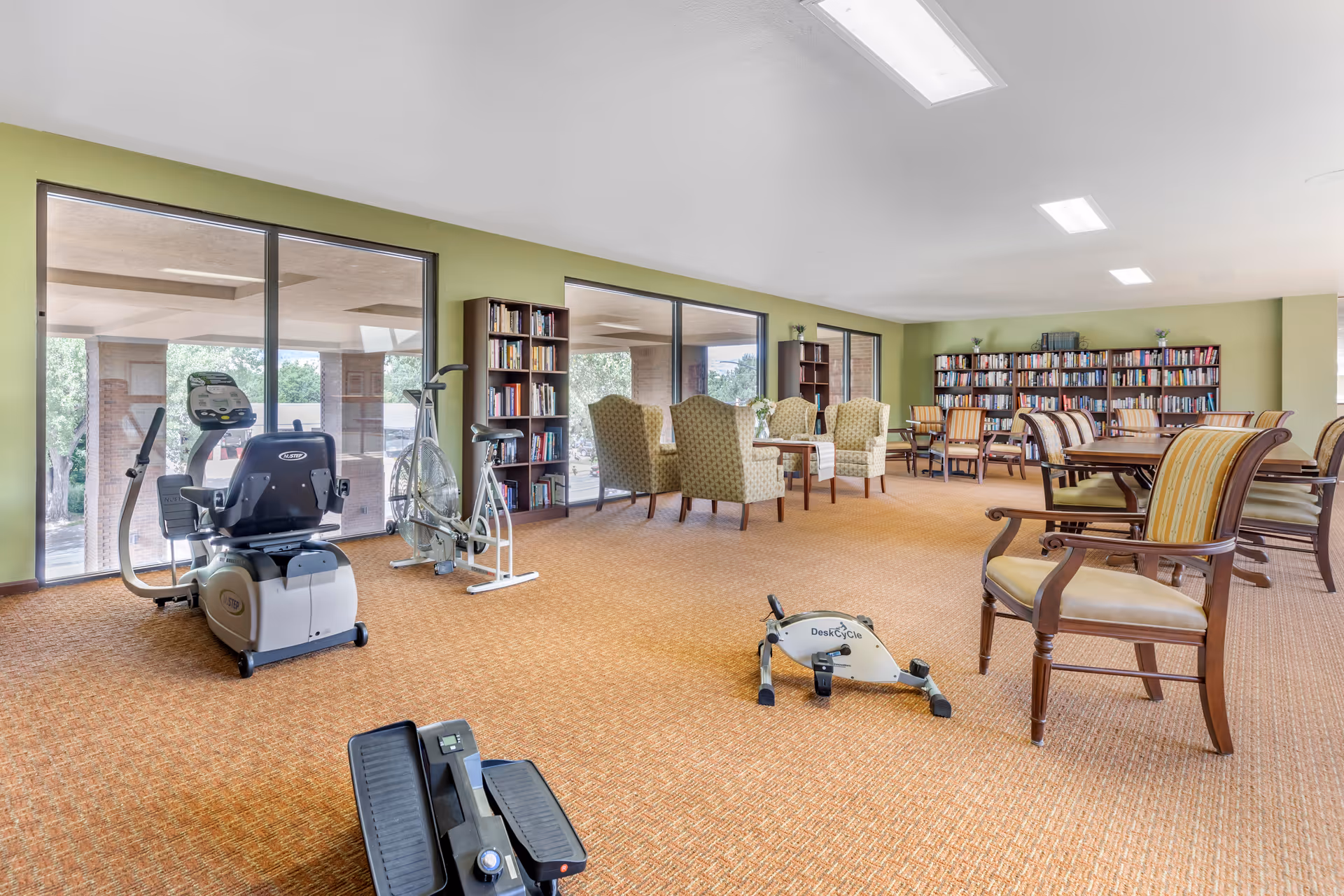 A spacious room with exercise equipment including a recumbent bike, an upright bike, and a DeskCycle pedal exerciser on a carpeted floor. The room also features several bookshelves filled with books, multiple upholstered armchairs, and tables arranged for seating. Large windows provide natural light and a view of the outdoor area.