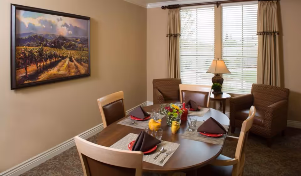 A dining room with a round wooden table set for four people with red plates, brown napkins, glasses, and silverware. There is a small flower arrangement and salt and pepper shakers in the center of the table. The room has beige walls, a large window with blinds and curtains, two brown upholstered armchairs, a small side table with a lamp and a plant, and a framed landscape painting on the wall.