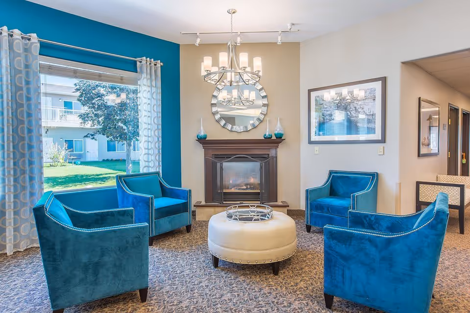 A cozy living room area with four blue velvet armchairs arranged around a round beige ottoman. There is a fireplace with a decorative round mirror above it and two vases on the mantel. A chandelier hangs from the ceiling, and a large window with patterned curtains shows a green outdoor area with trees and a building.