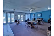 Community dining/activity room with several round tables and chairs, blue-painted walls, a ceiling fan, and large windows with vertical blinds.