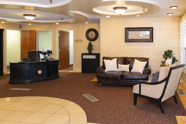 A cozy reception area with a dark wooden front desk on the left and a seating area on the right featuring a dark brown leather sofa with white pillows and a beige armchair. The walls are light-colored with a framed artwork and a round clock above a small black cabinet. The floor has a combination of beige tiles and brown carpet, and the ceiling has circular recessed lighting fixtures.