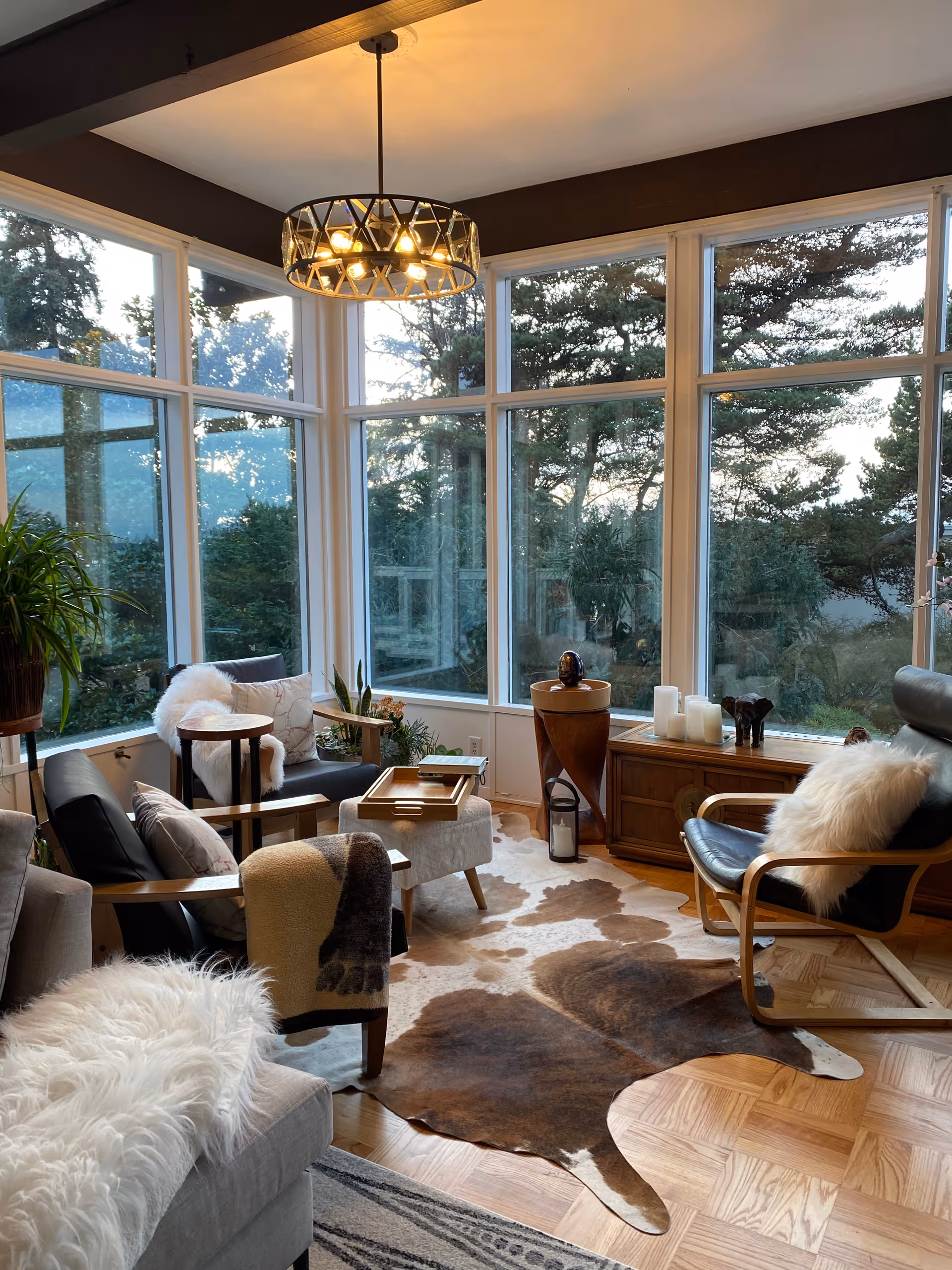 A cozy living room with large floor-to-ceiling windows showing a view of trees outside. The room features a mix of modern and rustic furniture including a gray sofa with a white fluffy throw, two armchairs with cushions and throws, a wooden coffee table, and a wooden sideboard with decorative items such as candles and a sculpture. A cowhide rug covers part of the wooden parquet floor, and a modern chandelier hangs from the ceiling.
